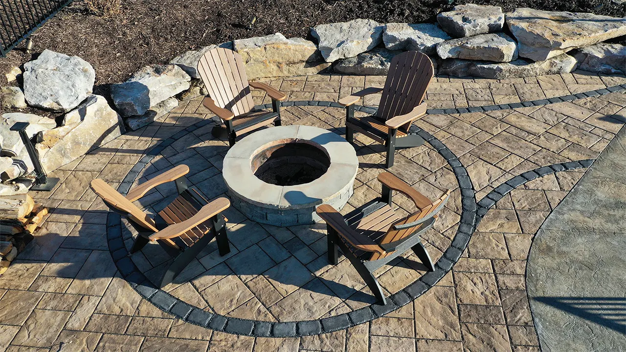 Paver patio with a round firepit, four chairs, a dark border inlay, and a boulder wall behind