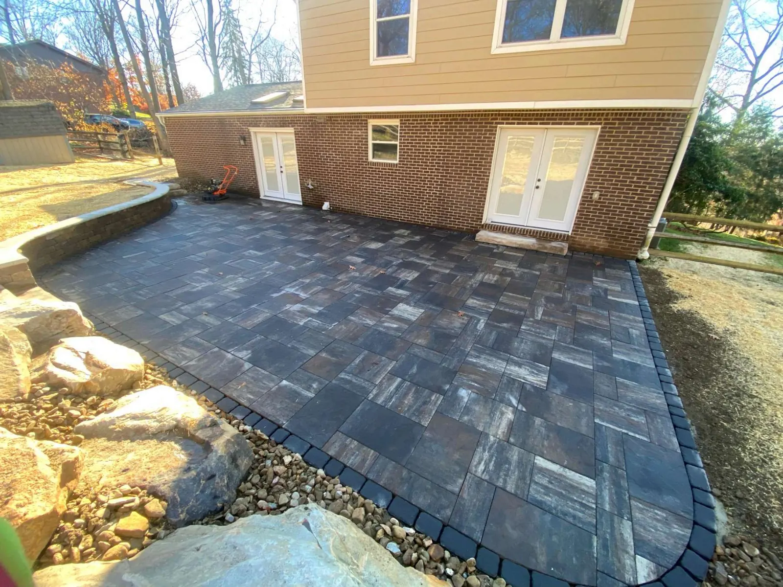 Extra large paver patio with a dark border beside boulders and a retaining wall