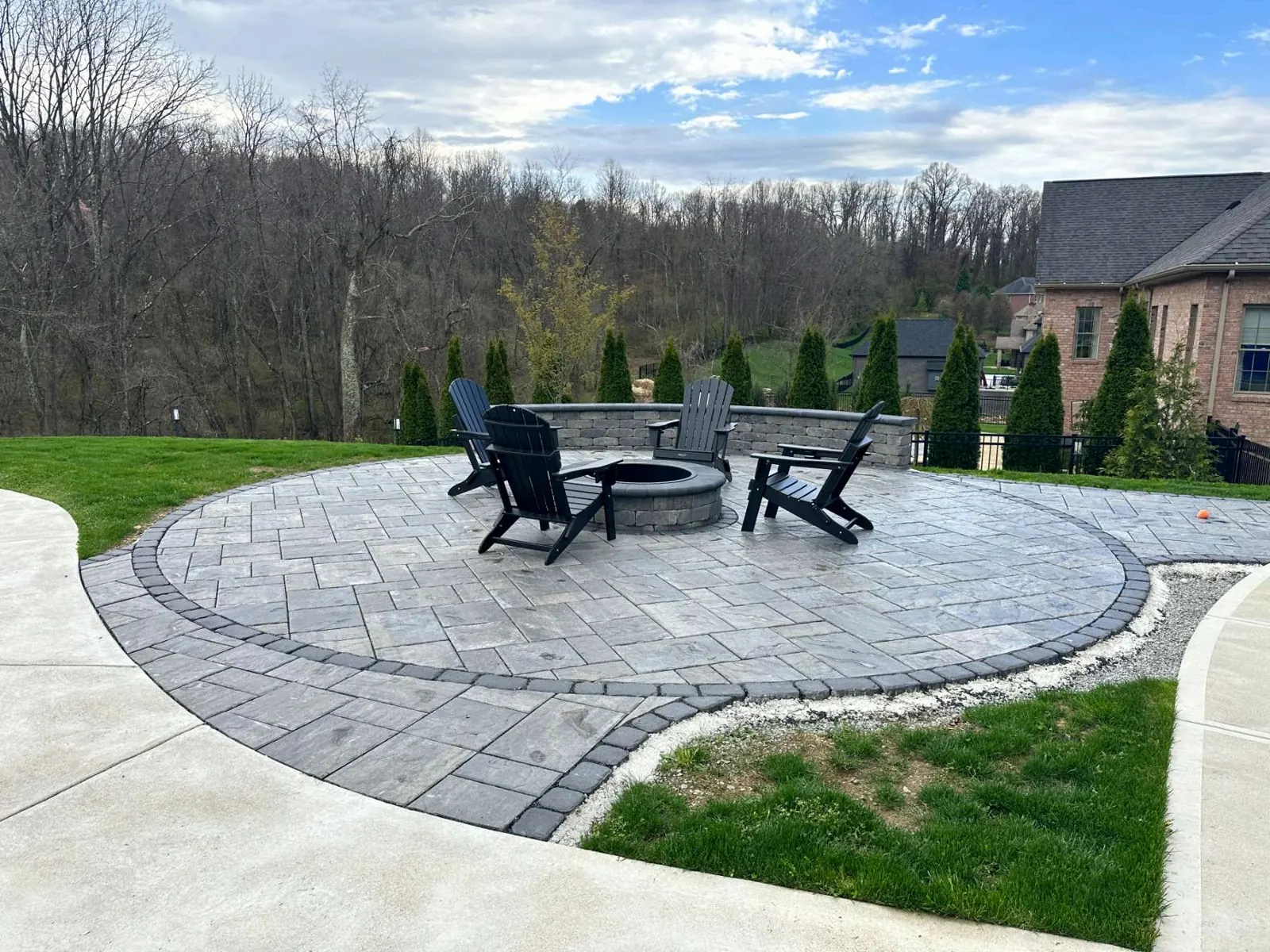 Large round paver patio with a firepit, chairs, and a stone wall behind