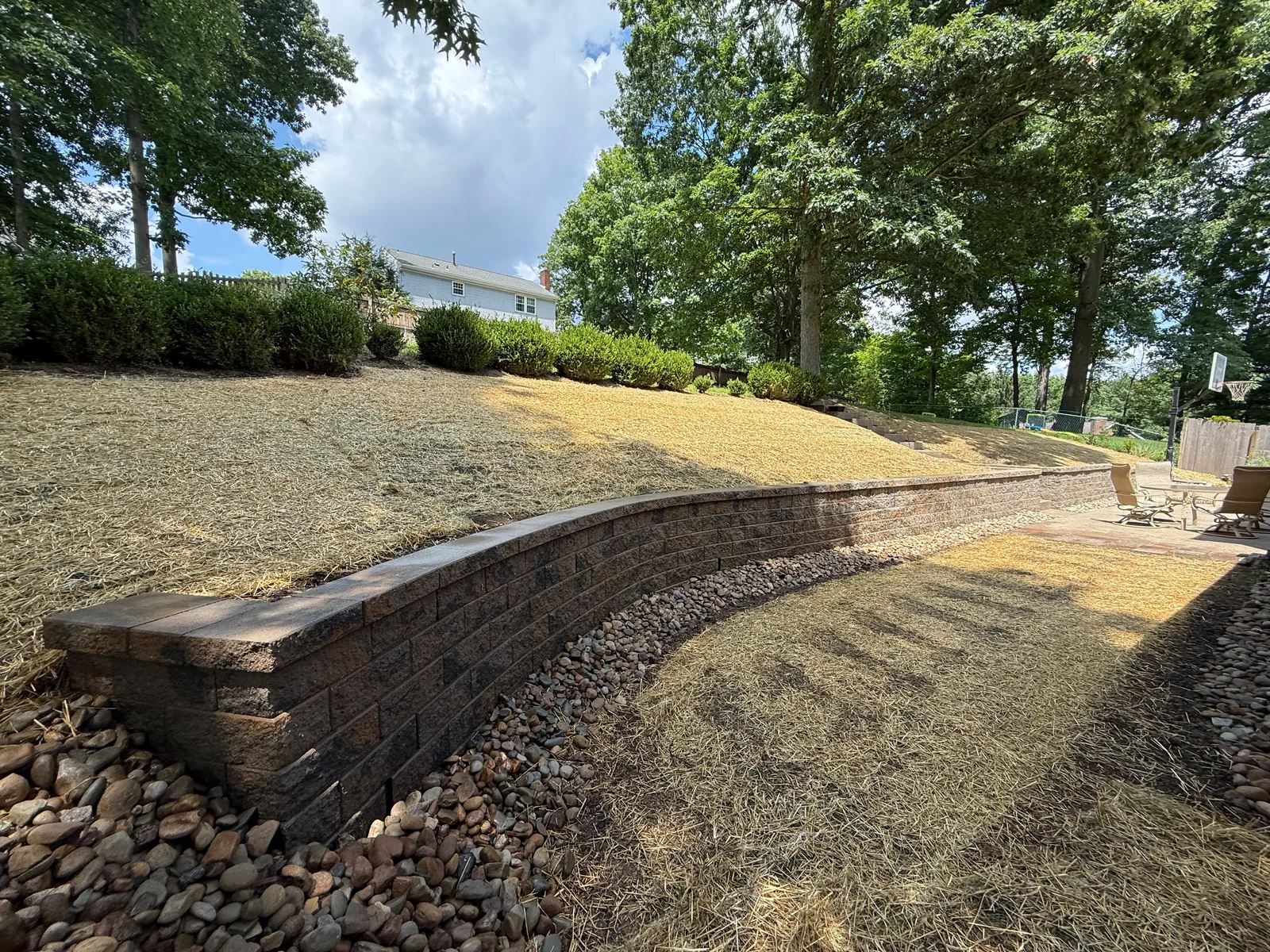 New curved Versa-Lok wall in the backyard with river rock along its base and straw over the slope