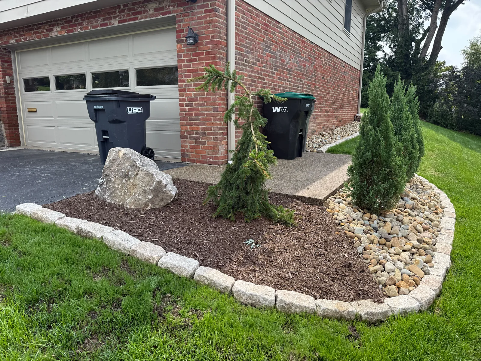 Mulch bed with a boulder, evergreens and river rock, edged with Belgian block, beside the garage