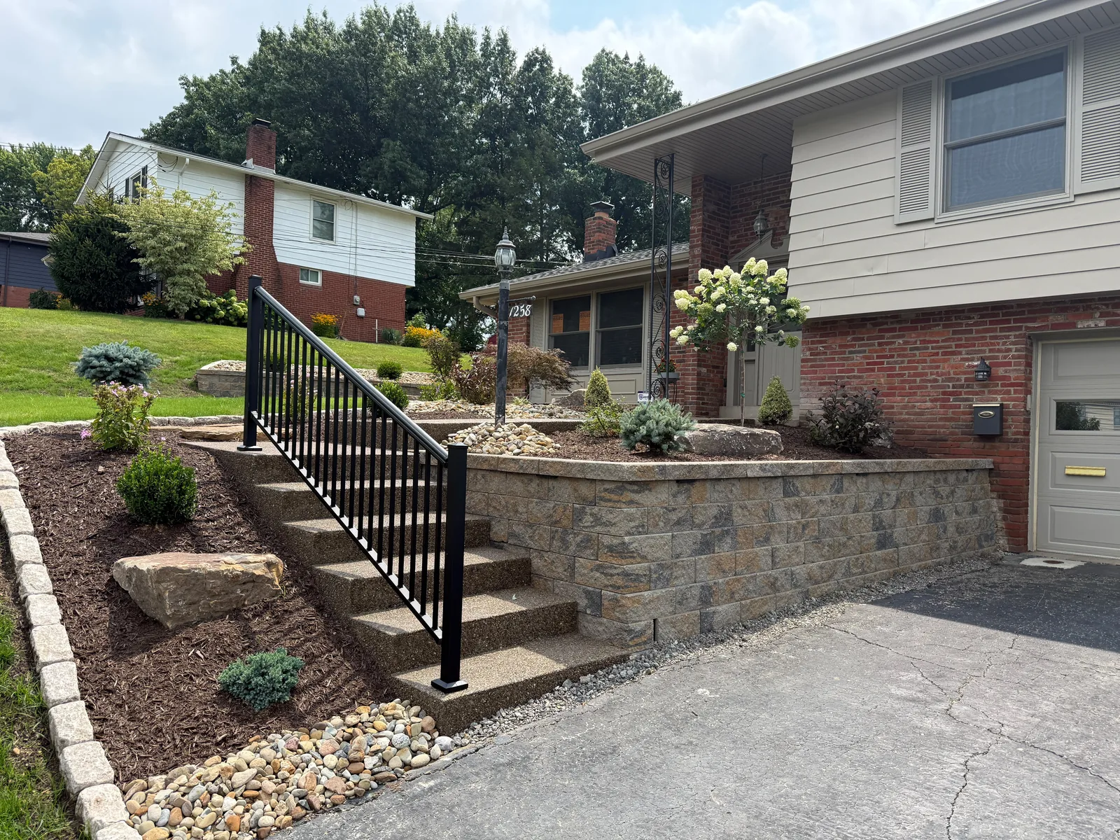 New front steps with a black railing, a Versa-Lok driveway wall, boulders, plantings and a Belgian block border at an Upper St. Clair home
